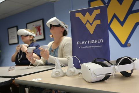 Photograph of people using virtual reality with Play Higher popup banner in front and Flying WV logo on the wall